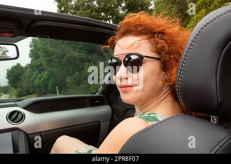 Porträt einer schönen jungen Frau mit Sonnenbrille, die in einem modernen Cabriolet sitzt. Porträt glückliche Frau im Cabrio. Reisen, Road Trip und People Concept - glückliche junge Frau im Cabrio Stockfoto