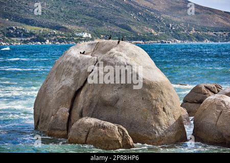 Große Felsen im blauen Ozean mit Bergen im Hintergrund. Atemberaubende Naturlandschaft oder Meereslandschaft an einem Sommertag. Felsbrocken oder große Natursteine im Meerwasser mit wunderschönen rauen Texturen Stockfoto