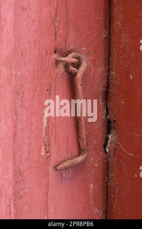 Vergrößerung der strukturierten roten Tür mit einem rostigen Metallhaken. Türrahmen und Türrahmen aus hellem Holz sind gerissen und weisen Risse, Kratzspuren und Holzsplitter auf. Außenansicht des Eingangs zum alten und abgenutzten Holzschuppen Stockfoto