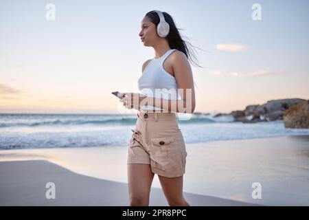 Nur ich, Musik und das Meer. Eine Frau, die Kopfhörer trägt und ihr Handy benutzt, während sie am Strand spazieren geht. Stockfoto