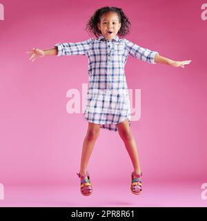 Ein hübsches, gemischtes Rennmädchen mit lockigem Haar feiert und tanzt vor einem pinkfarbenen Hintergrund in einem Studio. Afrikanisches Kind springt und fühlt sich frei Stockfoto