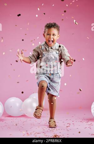 Ein süßer, gemischter Rennjunge, der vor einem rosa Hintergrund in einem Studio feiert und gewinnt. Ein afrikanisches Kind, das sich auf eine Party mit Konfetti und Ballons freut Stockfoto