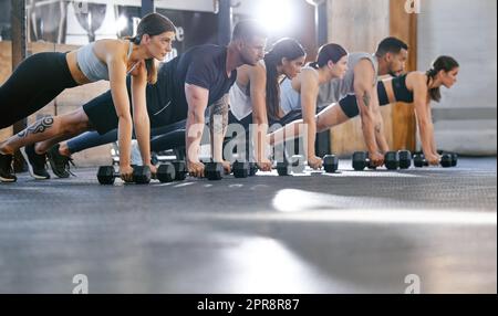 Eine vielfältige Gruppe aktiver junger Menschen, die beim Training in einem Fitnessraum gemeinsam an der Planke festhalten und Liegestützübungen mit Kurzhanteln machen. Fokussierte Athleten, die beim Workout in einem Fitnesskurs mit Gewichten auf Push-Ups und abtrünnigen Reihen trainieren, um Muskeln und Ausdauer aufzubauen Stockfoto