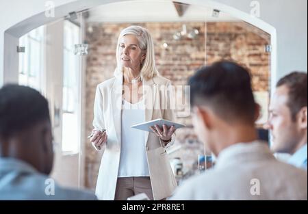 Wunderschöne, reife Architektin, die ein digitales Tablet verwendet, während sie bei einem Meeting im Vorstand mit ihren Kollegen und Kollegen spricht. Besprechung und Brainstorming von Plänen für ihr Entwicklungsprojekt Stockfoto