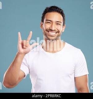 Ein hübscher junger Mann aus gemischten Rassen, der Rock-on-Rock-Sting, während er isoliert im Studio vor blauem Hintergrund stand. Hispanischer Mann, der die Gebärdensprache von "Ich liebe dich" zeigt, um Zuneigung oder Romantik zu zeigen Stockfoto