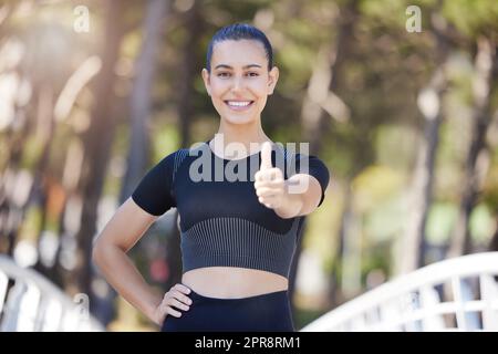 Glückliche Sportlerin, die beim Laufen oder Joggen im Freien Daumen nach oben zeigt. Fit, junge Frau, die beim Workout im Park lächelt und zufrieden aussieht Stockfoto