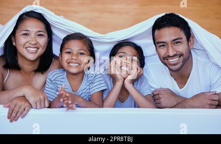 Glückliche Familie im Bett. Porträt einer hispanischen Familie im Bett. Junge Familie unter einem Bettlaken. Zwei Eltern, die sich mit ihren Töchtern verbinden.Schwestern, die sich mit ihren Eltern entspannen. Die Familie liegt im Bett Stockfoto