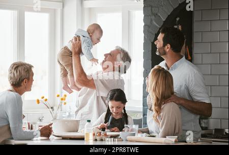 . Eine mehrgenerationale kaukasische Familienverbindung, während Sie den Tag zusammen am Küchentisch zu Hause verbringen. Großeltern, die das Neugeborene in der Hand halten. Ein junges Paar mit zwei Kindern. Stockfoto