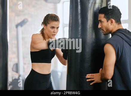 Passen Sie weiblichen Lernenden Boxen auf eine Boxtasche und holen Sie sich Tipps zum Fitnesstraining von ihrem Personal Trainer in einem Fitnessstudio. Starke Boxerin mit schlankem Körper, die mit ihrem Kampftrainer auf einen Kampf trainiert Stockfoto