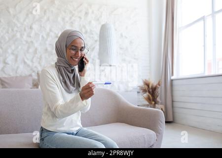 Glückliche junge muslimische Frau in Hidschab zu Hause, die zu Hause einen schnellen Schwangerschaftstest macht, ein positives Ergebnis bekam und Arzt, Ehemann rief. Stockfoto