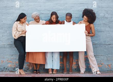 Gruppe von fünf verschiedenen jungen Geschäftsfrauen, die vor einer Mauer stehen und ein leeres Schild draußen in der Stadt sehen. Team von Kollegen, die ein Schild mit einer Nachricht halten, während sie gemeinsam im Freien in einer Schlange stehen Stockfoto