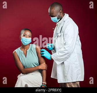 afroamerikanischer Arzt gibt Covid-Impfstoff an schwarze Frau, die chirurgische Gesichtsmaske trägt. Gesunder Patient erhält Corona-Injektion vom Arzt mit Nadel vor rotem Studiohintergrund mit Copyspace Stockfoto