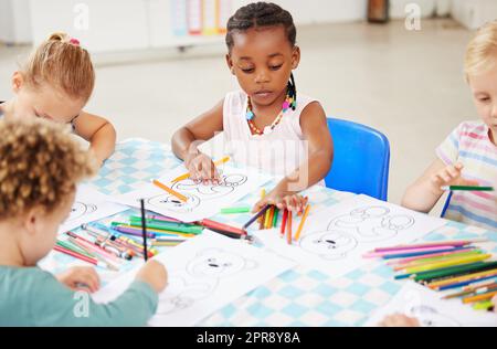 Diverse Gruppe von Kindern, die an einem Tisch sitzen und in der Vorschule oder im Kindergarten färben. Gruppe von Kindern mit bunten Stiften und Bildern Stockfoto