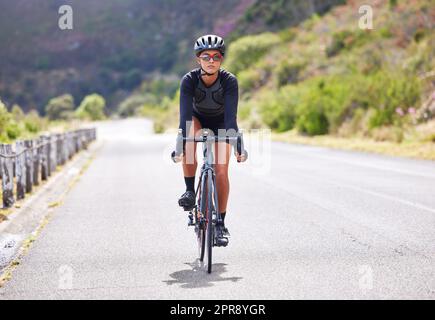 Eine sportliche junge Frau, die draußen radelt. Sportliche Passform für Frauen, die Helm und Brille tragen, während sie mit dem Fahrrad auf einer Straße entlang des Berges trainieren. Ausdauer und Kardio während eines Workouts und Trainings Stockfoto
