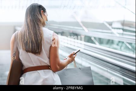 Rückansicht einer jungen Frau, die ein Handy benutzt, während sie auf einer Rolltreppe Einkaufstaschen in einem Einkaufszentrum trägt. Eine Frau, die gerne im Einzelhandel unterwegs ist und gleichzeitig mit ihrem Smartphone in Verbindung bleibt. Nutzen der App, um Angebote und Rabatte zu finden. Banking-App für bequemes Einkaufen Stockfoto