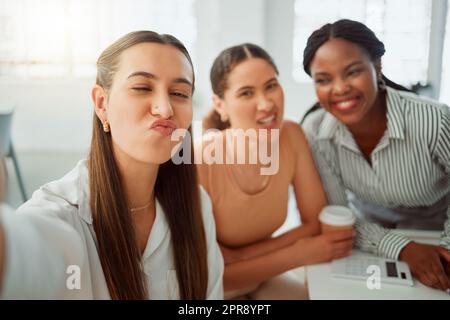 Porträt einer selbstbewussten jungen hispanischen Geschäftsfrau, die einen lustigen Gesichtsausdruck macht, während sie mit ihren Kollegen in einem Büro Selfies macht. Eine Gruppe von drei glücklichen lächelnden Frauen, die als engagiertes und ehrgeiziges Team in einer kreativen Startup-Agentur Fotos machen Stockfoto