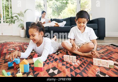 Kleine Kinder, die auf dem Boden sitzen, mit Spielzeug und Farben in einem Buch. Kleine gemischte Geschwister spielen zu Hause zusammen, während die Eltern im Hintergrund sitzen Stockfoto