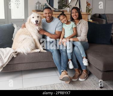 Porträt einer glücklichen Mischfamilie von drei Personen, die sich mit ihrem Hund auf dem Sofa entspannen. Liebende schwarze Familie, die liebevoll mit einem Pflegekind ist. Ein junges Paar, das sich mit seiner Tochter anfreundet und das Welpe zu Hause gerettet hat Stockfoto