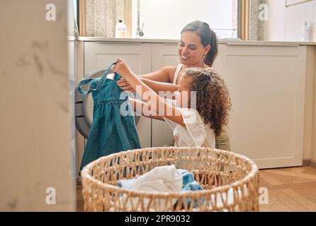 Junge hispanische Mutter und ihre Tochter sortieren schmutzige Wäsche in der Waschmaschine zu Hause. Ein bezauberndes kleines Mädchen, das seiner Mutter bei Hausarbeiten hilft Stockfoto