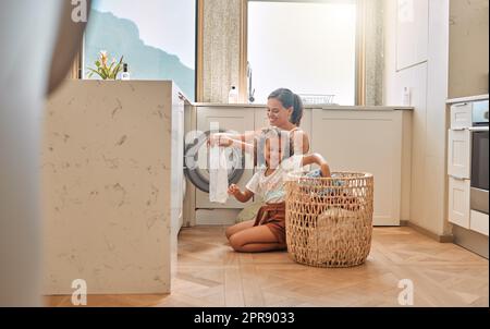 Junge hispanische Mutter und ihre Tochter sortieren schmutzige Wäsche in der Waschmaschine zu Hause. Ein bezauberndes kleines Mädchen und ihre Mutter machen Hausarbeiten zusammen Stockfoto