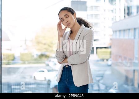 Junge unglückliche gemischte Geschäftsfrau, die unter Kopfschmerzen leidet, wenn sie im Büro steht. Ein müder hispanischer Geschäftsmann, der an einem Fenster auf der Arbeit stand und einen Burn-out hatte Stockfoto