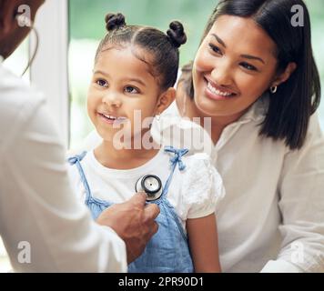 Doktor untersucht glückliches kleines Mädchen mit Stethoskop. Kind sitzt mit Mutter, während männlicher Kinderarzt auf Herzschlag hört. Mutter hält Tochter während Arztbesuch Stockfoto