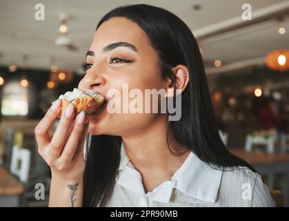 Die perfekte Zeit zum Genießen. Eine schöne junge Frau, die in einem Café zu einem leckeren Leckerbissen beißt. Stockfoto