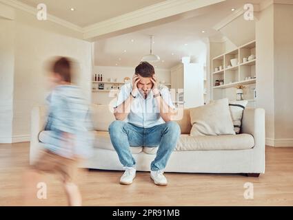 Junger, weißer Vater, der Kopfschmerzen hat, während sein Sohn zu Hause in der Lounge läuft. Ein sorgenfreier Junge, der im Wohnzimmer rennt, während sein Vater müde aussieht. Ein Mann, der aufgebracht aussieht, während sein Kind spielt Stockfoto