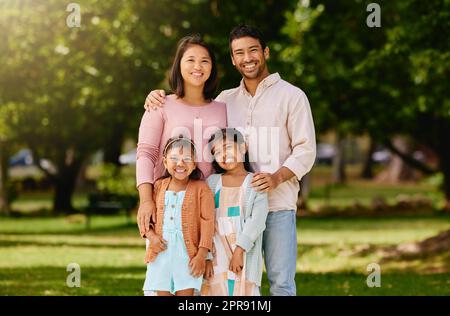 Porträt einer glücklichen Mischrassenfamilie, die an einem sonnigen Tag in einem öffentlichen Park zusammenstand. Asiatische Eltern und zwei kleine Töchter genießen einen Tag im Freien Stockfoto