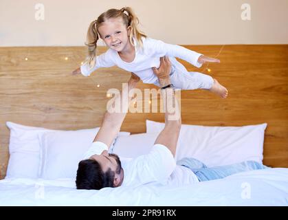 Ein bezauberndes kleines Mädchen, das sich mit ihrem alleinerziehenden Vater zu Hause anfreundet und so tut, als könne sie fliegen. Alleinerziehende kaukasische Eltern, die ihre Tochter in der Luft halten und heben. Lächelndes Kind, das im Schlafzimmer spielt Stockfoto