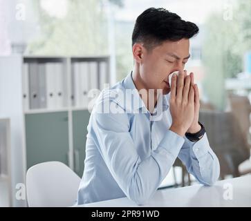 Ein kranker Geschäftsmann, der ein Taschentuch benutzt und sich die Nase putzt, während er in einem modernen Büro arbeitet. Ein kranker asiatischer Mann erkrankt an Grippe, einer Erkältung oder einem Fleck. Männlicher Angestellter sitzt am Tisch und niest, fühlt sich unglücklich Stockfoto