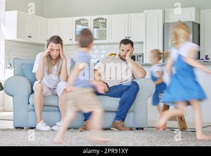 Gestresste Eltern, die auf der Couch sitzen und Kinder herumlaufen. Aufgeregte Kinder, die um verärgerte Eltern spielen. Frustrierte Kinder, die herumrennen. Unglückliche Eltern, die sich über Kinder aufregen. Kinder spielen Stockfoto