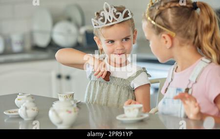 Zwei kleine Mädchen feiern zu Hause eine Prinzessinnen-Teeparty. Geschwisterschwestern tragen Diademe, während sie mit dem Teeset spielen und Kekse am Küchentisch essen. Schwestern verstehen sich gut und spielen zusammen Stockfoto