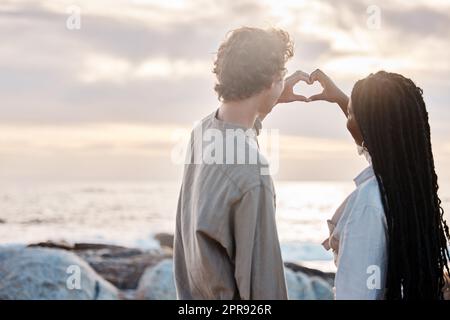 Rückansicht eines jungen multirassischen Paares, das romantisch ist, indem es mit den Händen am Strand eine Herzgeste macht Stockfoto