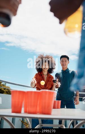 Fröhliche junge Freunde spielen draußen Bier-Pong. Verschiedene Freunde werfen einen Ball in Bierbecher. Junger asiatischer Mann, der Spaß hat und ein Spiel mit seinem lateinamerikanischen Freund spielt. Lächelnde junge Freunde Stockfoto