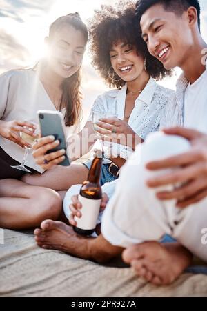 Eine Gruppe von drei multirassischen Freunden, die sich am Strand entspannen und mit einem Telefon telefonieren, während sie alkoholische Getränke trinken Stockfoto