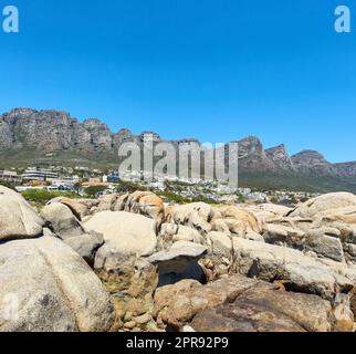 Zwölf Apostel am Tafelberg in Kapstadt vor einem klaren blauen Himmelshintergrund an einem sonnigen Tag mit Kopierraum. Blick auf einen ruhigen Vorort, umgeben von malerischer Berglandschaft und Strandfelsen Stockfoto