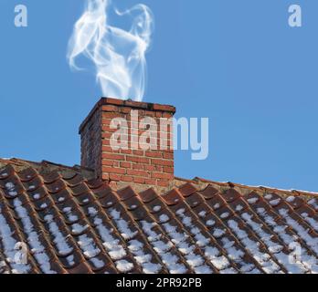 Im Winter bläst der Schornstein Rauch auf die Dachterrasse des Hauses in Dänemark. Nahaufnahme der alten Architektur mit rotem Backsteinluftschacht, um Hitze und Rauch vom Kamin mit blauem Himmel zu entfernen Stockfoto