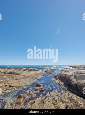Kopieren Sie Platz auf See mit einem klaren blauen Himmelshintergrund und felsiger Küste in Camps Bay, Kapstadt, Südafrika. Felsbrocken an einem Strandufer über einem majestätischen Ozean. Malerische Landschaft für einen Sommerurlaub Stockfoto