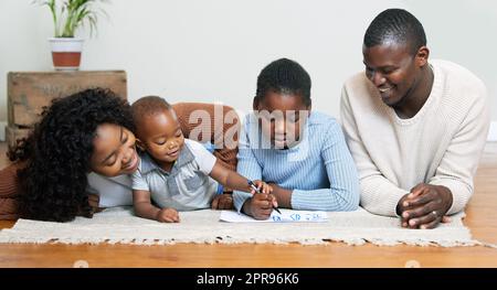 Ich helfe Ihnen. Zwei junge Eltern, die mit ihren Kindern auf dem Boden des Wohnzimmers liegen und ihnen beim Zeichnen zusehen. Stockfoto