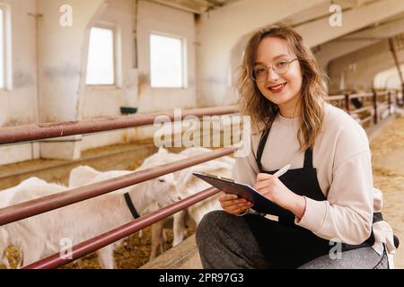 Junge lächelnde Landwirtin in einer Schürze mit Tablet auf einer Ziegenfarm. Milchproduktion. Konzept für kleine Unternehmen. Stockfoto