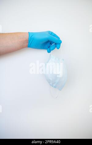 Eine Hand in blauen Handschuhen trägt eine weiße medizinische Schutzmaske auf weißem Hintergrund. Stockfoto