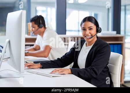 Überlassen Sie uns die Lösung von Problemen. Zwei junge Geschäftsleute, die zusammen im Büro sitzen und Headsets tragen, während sie Computer benutzen. Stockfoto