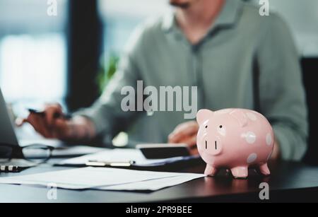 Versuchen Sie nicht, das Sparschwein zu brechen, wenn Sie Geld ausgeben. Ein unkenntlich Geschäftsmann, der Papierkram mit einem Sparschwein auf seinem Schreibtisch bei der Arbeit macht. Stockfoto