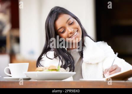 Beginnen Sie Ihren Tag immer mit einer guten Note. Eine junge Frau, die beim Brunch in einem Notizbuch schreibt. Stockfoto