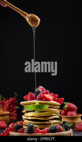 Stapel von Pfannkuchen mit frischem Obst bestreut mit Honig auf schwarzem Hintergrund Stockfoto