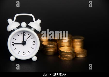 Weißer Wecker mit Goldmünzen, isoliert auf dunklem Hintergrund. Kopierbereich und Geschäftskonzept Stockfoto