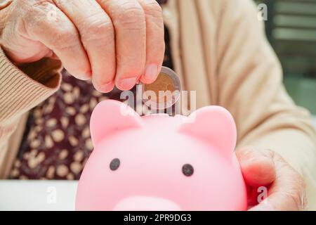 Ältere Frau im Ruhestand, die Münzen auf Sparschwein legt und sich um die monatlichen Ausgaben und die Bezahlung der Behandlungsgebühr sorgt. Stockfoto