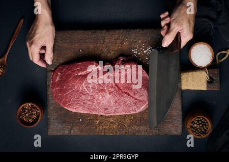 Rohes Rinderfilet liegt auf einem hölzernen Schneidebrett und Gewürze zum Kochen auf einem schwarzen Tisch, Draufsicht. Die Hand einer Frau hält ein großes Küchenmesser in der Hand Stockfoto