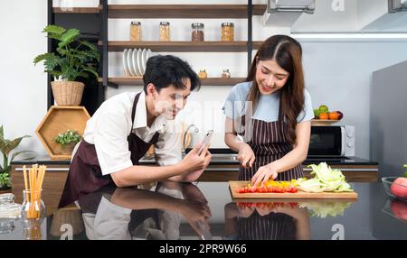 Asiatisches Paar verbringt Zeit zusammen in der Küche. Junge Frau in Schürze kocht Salatgericht, während sein Freund ein Video für einen sozialen Blogger aufnimmt. Moderner Lebensstil, Beziehung und Aktivität Stockfoto